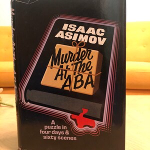 May include: A hardcover book titled "Murder at the ABA" by Isaac Asimov. The cover is black with a graphic featuring a wrapped package and the title in a retro font. The book's tagline reads "A puzzle in four days & sixty scenes."