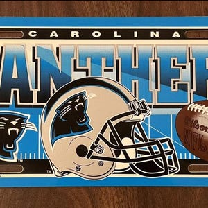 May include: Carolina Panthers license plate with the team name and logo. The plate features a black panther logo, a football helmet, and a football. The background is blue and white.