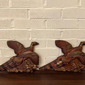 May include: Two decorative pheasant wall sculptures. The birds are in a resting pose with their wings spread. The sculptures are brown with bronze and gold accents. The background is a white brick wall.