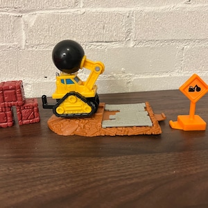 May include: A yellow toy construction vehicle with a black wrecking ball, a gray and brown playset, and three red toy blocks. The vehicle is on the playset and the blocks are stacked on the wooden surface.