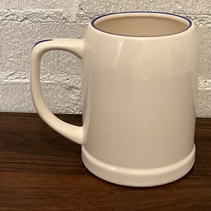 May include: A white ceramic beer stein with a rounded body and a sturdy handle. The rim and handle are accented with a thin blue line. The mug sits on a dark wooden surface, with a white brick wall in the background.