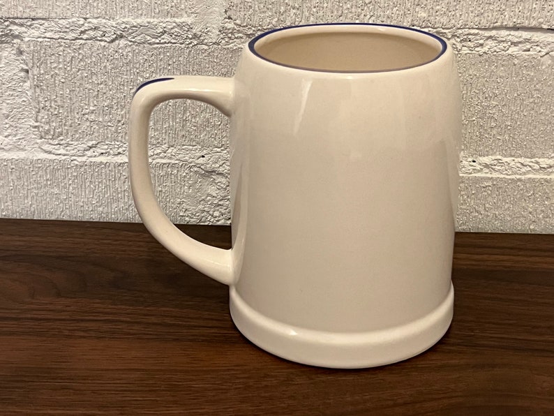 May include: A white ceramic beer stein with a rounded body and a sturdy handle. The rim and handle are accented with a thin blue line. The mug sits on a dark wooden surface, with a white brick wall in the background.