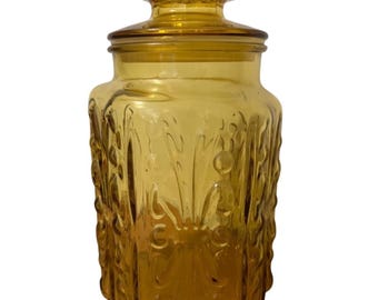 May include: Amber glass jar with a lid, featuring a raised floral design. The jar is square shaped and has a textured surface.