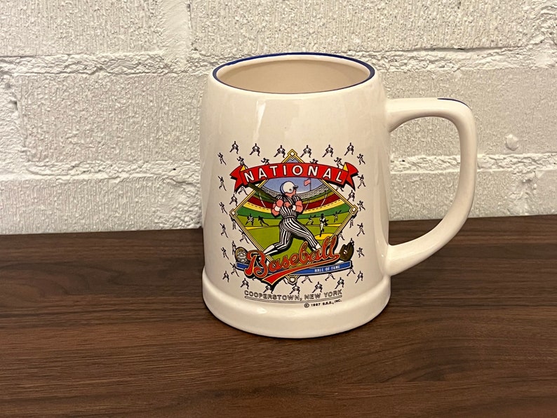 May include: Cream-colored ceramic beer stein with a blue rim and a large handle. The stein features a baseball-themed graphic with the words "National Baseball Hall of Fame" and "Cooperstown, New York" printed on it.