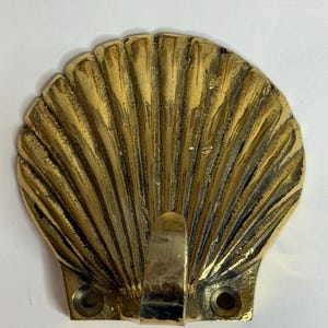 May include: A polished brass wall hook shaped like a scallop shell. The hook features a fan-like design with vertical ridges radiating from the base. Two circular mounting holes are positioned at the bottom corners for secure installation.