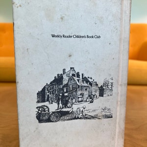 K&ouml;nnte beinhalten: Ein wei&szlig;es Buch mit schwarzem Text, der "Weekly Reader Children's Book Club" lautet. Der hintere Umschlag zeigt eine Schwarzwei&szlig;illustration einer Stra&szlig;enszene in der Stadt mit Geb&auml;uden, Menschen und einem Schiff.