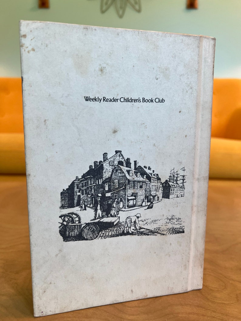 K&ouml;nnte beinhalten: Ein wei&szlig;es Buch mit schwarzem Text, der "Weekly Reader Children's Book Club" lautet. Der hintere Umschlag zeigt eine Schwarzwei&szlig;illustration einer Stra&szlig;enszene in der Stadt mit Geb&auml;uden, Menschen und einem Schiff.
