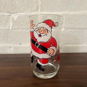 May include: Clear glass with a cartoon Santa Claus design. Santa is depicted in his traditional red suit, black belt, and white beard, waving with a smile. The glass is cylindrical with a slightly flared base, suitable for beverages.