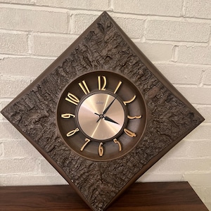 May include: Vintage brown wall clock with a diamond-shaped, textured frame. The clock face is gold with gold numerals and hands. The clock is mounted on a white brick wall and a dark brown wooden surface.