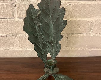 May include: A bronze-colored candle holder shaped like oak leaves. The candle holder has a small cup for a candle and a base with three leaf-shaped feet. The leaves have detailed veining and a textured surface.