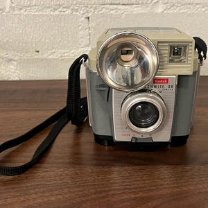May include: Vintage Kodak 20 camera with a silver and gray body, a large flash, and a black strap. The camera has a lens and a viewfinder. The camera is sitting on a wooden surface.