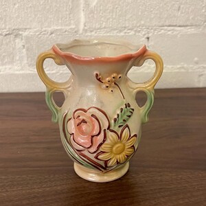 May include: A small, cream-colored vase with two handles and floral designs. The vase features a pink rose and a yellow sunflower, with green leaves and accents. The rim is a peach color, and the handles are yellow and green.