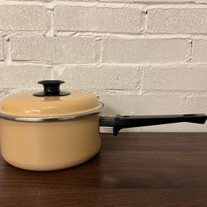May include: A beige enamel saucepan with a black handle. The saucepan has a silver rim around the top.