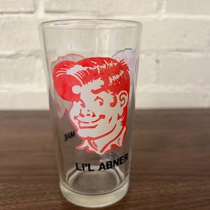 May include: A clear glass drinking glass with a red and white cartoon image of a man with the text "Li'l Abner" printed on the bottom.