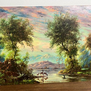 May include: A vintage landscape painting depicts a serene scene with a lake, trees, and mountains under a colorful sky. Deer are drinking from the water. Birds fly overhead, adding to the tranquil atmosphere. The artwork is framed.