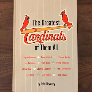 May include: A book cover with the title "The Greatest Cardinals of Them All." The cover features a red and orange logo with two cardinal birds. The names of baseball players are listed below the title.