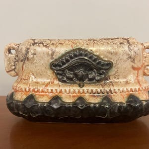 May include: A rectangular ceramic planter with a cream-colored, textured surface and a decorative black and bronze design. The planter has two handles and a black, scalloped base. The planter is suitable for indoor or outdoor use.