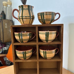May include: Set of six brown and green ceramic mugs with a plaid pattern, displayed in a wooden shelf unit.