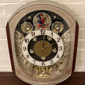 May include: A Seiko wall clock with a champagne-colored frame and dark wood side accents. The clock face features black Arabic numerals, a central gold dial, and two dancing figures in top hats. Decorative crystal elements and a pendulum are visible.