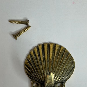 May include: A brass wall hook shaped like a scallop shell with a textured, ribbed surface and a central hook protrusion. The base features two circular mounting holes for installation, and two matching brass screws are included for mounting.