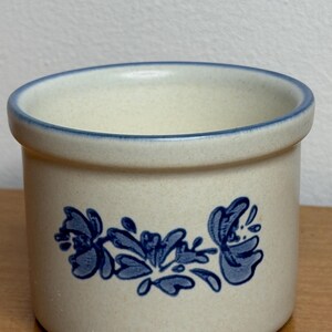 May include: A cylindrical stoneware crock with a speckled off-white finish and a blue rim. The exterior features a blue floral pattern centered on the front. The vessel has a thick, rounded top edge and a flat base, providing a sturdy structure.