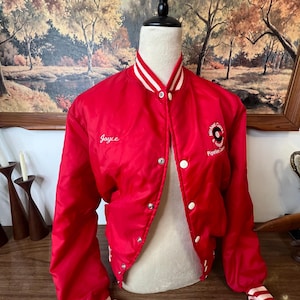 May include: A red satin bomber jacket with white accents. The jacket has a white collar and cuffs with white stripes. The name "Joyce" is embroidered on the front, and a circular logo is on the right side. The jacket is buttoned up.