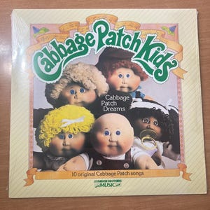 May include: A vintage Cabbage Patch Kids album cover featuring five dolls with different hair colors and styles. The album is titled "Cabbage Patch Dreams" and includes "10 original Cabbage Patch songs". The Parker Brothers Music logo is visible.