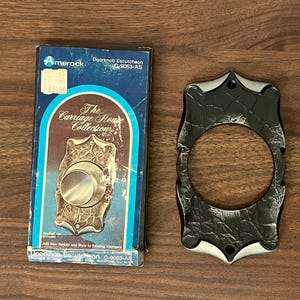 May include: A black and silver door knob escutcheon with a decorative design. The escutcheon is labeled "The Carriage House Collection" and "Amerock Doorknob Escutcheon C-9063-AS".