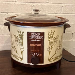 May include: A vintage Hamilton Beach Crock Watcher slow cooker with a cream-colored exterior, brown botanical illustrations, and a glass lid. The front control panel features the text Crock Watcher, 6 Quart Cook'n Serve Removable Vessel, and Automatic Shift with settings for Off, Low, High, and Auto Shift.