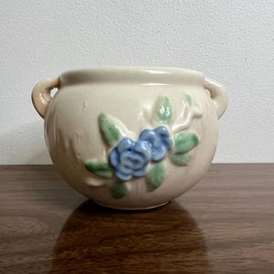May include: A white ceramic planter with blue and green floral designs. The planter has two handles and is a round shape.