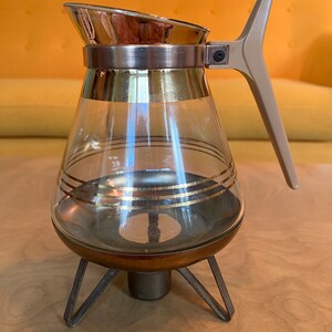 May include: A glass coffee pot with a gold trim and a brown handle. The pot sits on a metal stand with three legs.
