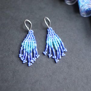 Peut inclure: Paire de boucles d'oreilles faites à la main, composées de perles bleues et bleu clair. Les boucles d'oreilles ont une forme de goutte d'eau avec une frange de perles. Elles sont posées sur une surface gris foncé, avec des flacons de perles bleues en arrière-plan.