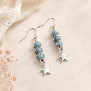 Peut inclure: Une paire de boucles d'oreilles pendantes composées de trois perles rondes bleu gris entourées de fil argenté. Chaque boucle d'oreille est suspendue à un crochet argenté et se termine par une petite breloque étoile à cinq branches en métal argenté.