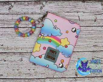 May include: A pink wallet with a rainbow, clouds, and stars design, with the text "Yari's Stitchy Station". A colorful coil bracelet is next to the wallet. The wallet has a pink border and silver snaps.