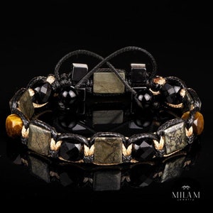 Puede incluir: Un brazalete trenzado negro con cuentas cuadradas verdes y doradas, cuentas redondas negras y cuentas de ojo de tigre.