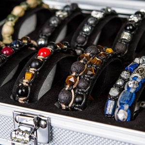 May include: A silver metal case with a black interior holds a collection of beaded bracelets. The bracelets are made with various materials, including lava rock, tiger's eye, and lapis lazuli.