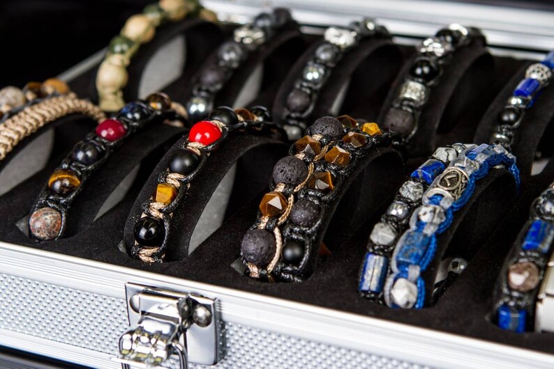 May include: A silver metal case with a black interior holds a collection of beaded bracelets. The bracelets are made with various materials, including lava rock, tiger's eye, and lapis lazuli.