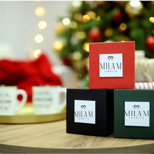 May include: Three small gift boxes with the logo "MILAM jewelry" on each. The boxes are red, black, and green. The boxes are sitting on a wooden table.