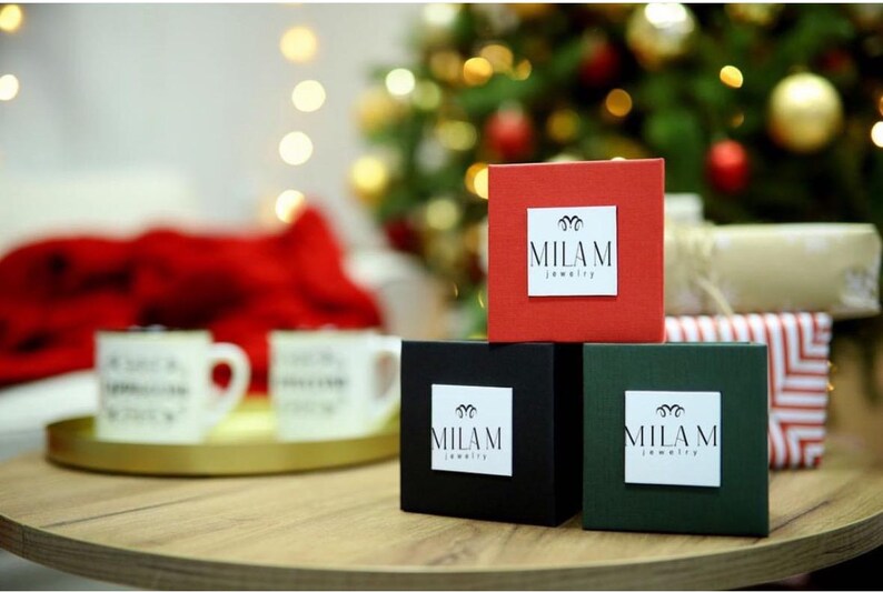 May include: Three small gift boxes with the logo "MILAM jewelry" on each. The boxes are red, black, and green. The boxes are sitting on a wooden table.