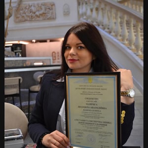 May include: A woman holds a framed certificate with the text "СВІДОЦТВО CERTIFICATE МАМРИГА ЛЮДМИЛА АНАТОЛІЇВНА MAMBYMA LIUOMYLA АТЕСТАЦІЯ ТА ЕКСПЕРТНА ОЦІНКА ДОРОГОЦІННОГО КАМІННЯ" in Ukrainian.
