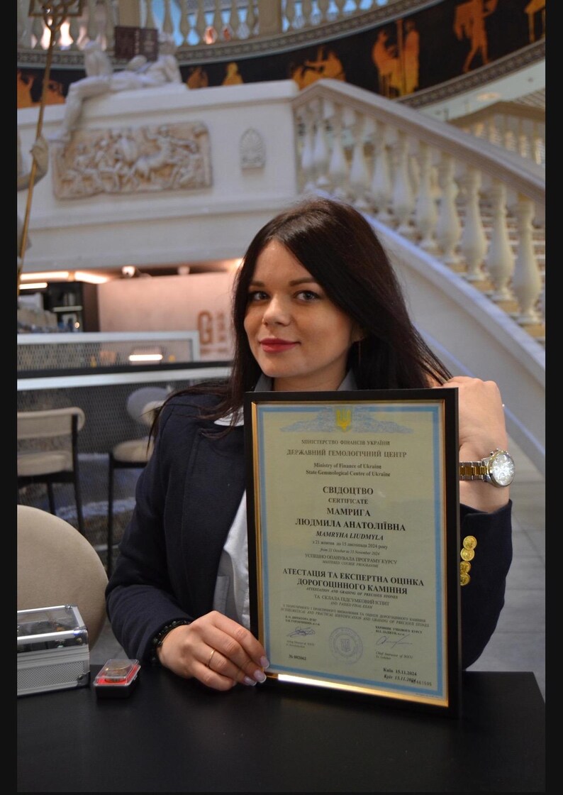 May include: A woman holds a framed certificate with the text "СВІДОЦТВО CERTIFICATE МАМРИГА ЛЮДМИЛА АНАТОЛІЇВНА MAMBYMA LIUOMYLA АТЕСТАЦІЯ ТА ЕКСПЕРТНА ОЦІНКА ДОРОГОЦІННОГО КАМІННЯ" in Ukrainian.