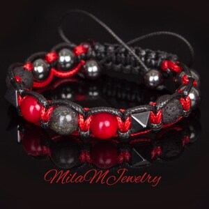 May include: A red and black braided bracelet with red, black, and grey beads. The bracelet has a black cord with a knot at the end. The bracelet is on a black background. Mila M Jewelry.