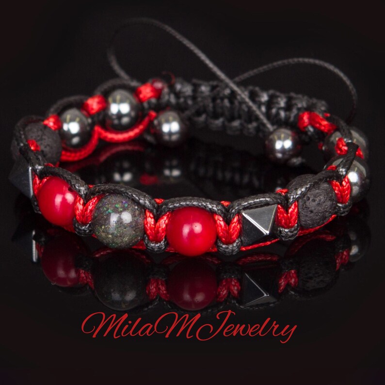 May include: A red and black braided bracelet with red, black, and grey beads. The bracelet has a black cord with a knot at the end. The bracelet is on a black background. Mila M Jewelry.