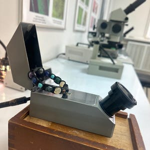 May include: A gray refractometer with a black lens, resting on a wooden box. A colorful beaded bracelet is wrapped around the refractometer.