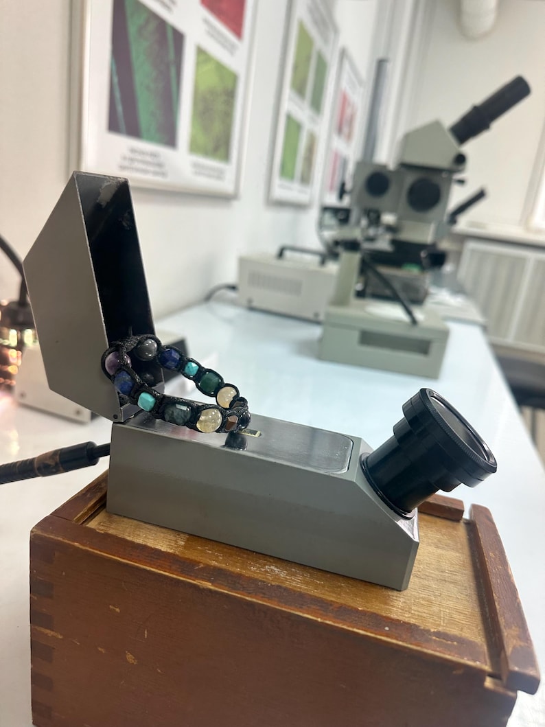 May include: A gray refractometer with a black lens, resting on a wooden box. A colorful beaded bracelet is wrapped around the refractometer.