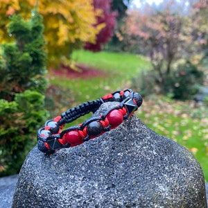 May include: A black beaded bracelet with red gemstone beads and black lava beads. The bracelet is tied with a black cord and has a knot closure.