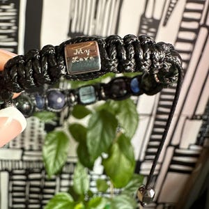 May include: Black braided bracelet with a silver charm that says "My Love". The bracelet also has blue and green gemstone beads.