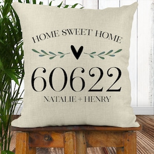 May include: A beige throw pillow with the text "HOME SWEET HOME" in black lettering, a black heart, and green leafy branches. The number "60622" is printed in black below the text, and the names "NATALIE + HENRY" are printed in black below the number.