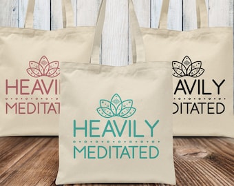 May include: Three canvas tote bags with the text "Heavily Meditated" printed on them. The bags are beige with black handles. The text is in different colors on each bag: pink, teal, and black.
