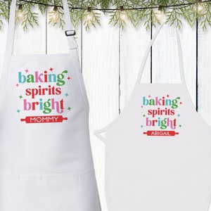May include: Two white aprons with the text "baking spirits bright" in colorful letters. The larger apron says "Mommy" below the text, and the smaller apron says "Abigail". Both aprons have a red rolling pin graphic.